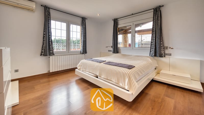 Villas de vacances Costa Brava Espagne - Villa Soleia - Chambre avec un lit double blanc, parquet en bois et grandes fenêtres habillées de rideaux gris à motifs.