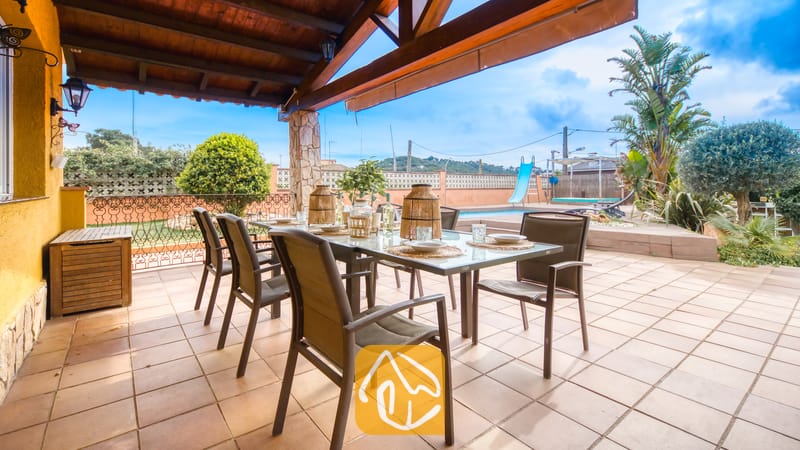 Villas de vacances Costa Brava Espagne - Villa Soleia - Terrasse extérieure couverte de la Villa Soleia dotée d'une table avec plateau en verre, de chaises en métal foncé, de lanternes de table tressées et d'une vue sur la piscine et le jardin.