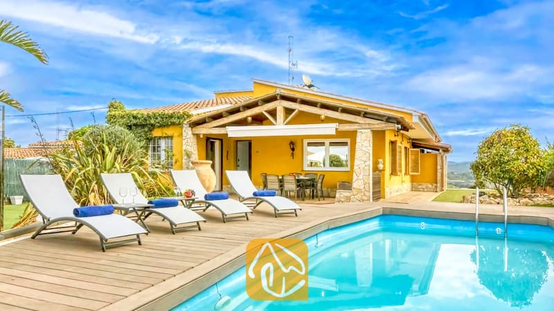 Vakantievilla's Costa Brava Spanje - Villa Soleia - Privézwembad met houten vlonder en witte ligstoelen bij Villa Soleia in Spanje, met een villa met een gele buitenkant en een blauwe lucht.