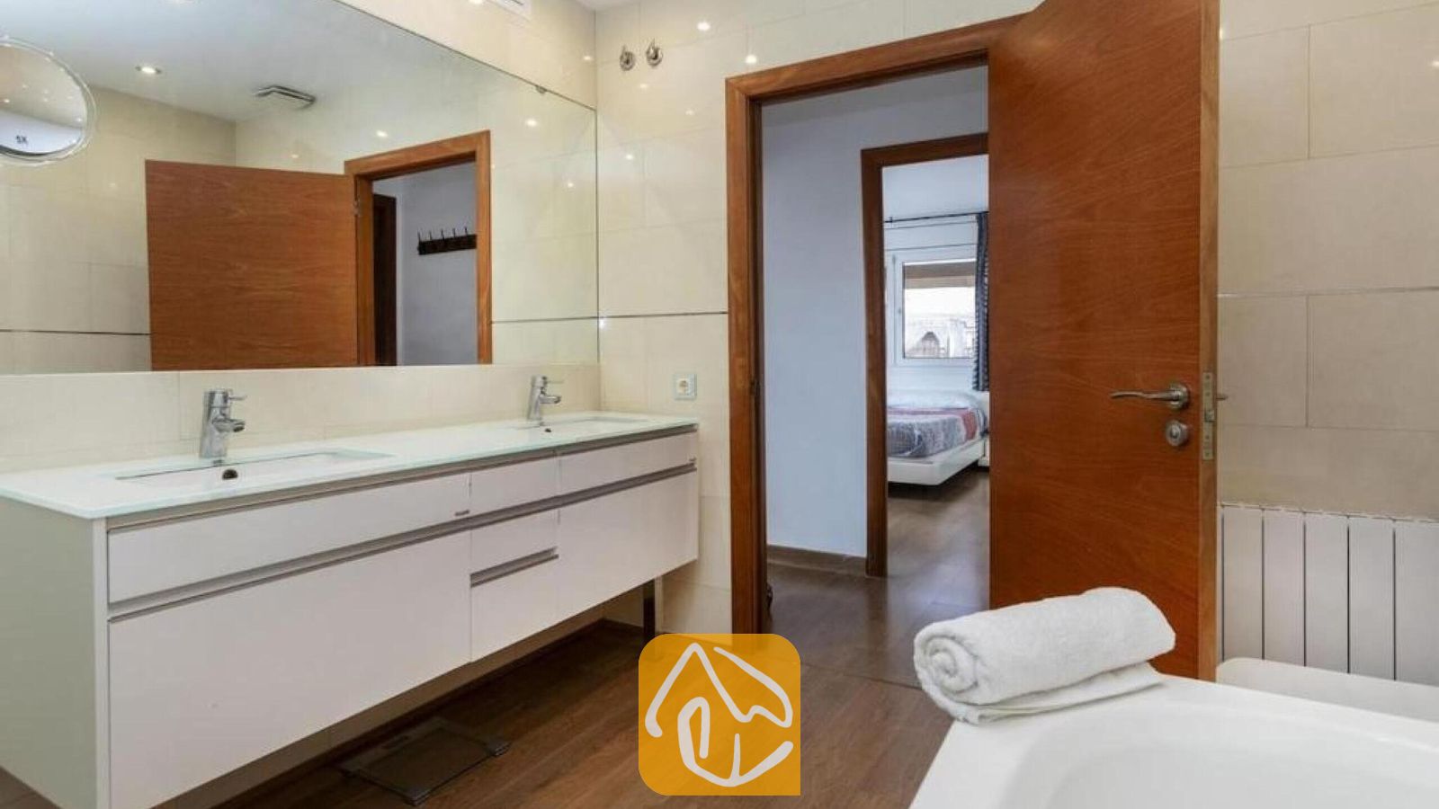 Vakantievilla's Costa Brava Spanje - Villa Soleia - Badkamer voorzien van een wit dubbel meubel met twee wastafels, een grote wandspiegel en een houten deur die toegang biedt tot een aangrenzende slaapkamer.