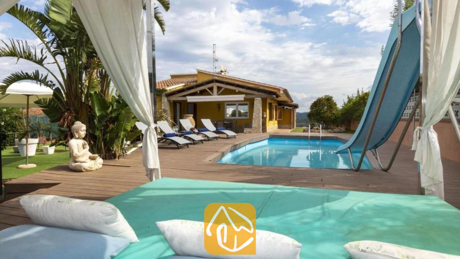 Vakantievilla's Costa Brava Spanje - Villa Soleia - Buitenzwembad en houten terras bij Villa Soleia, voorzien van ligstoelen, een blauwe waterglijbaan en een geel huis op de achtergrond.