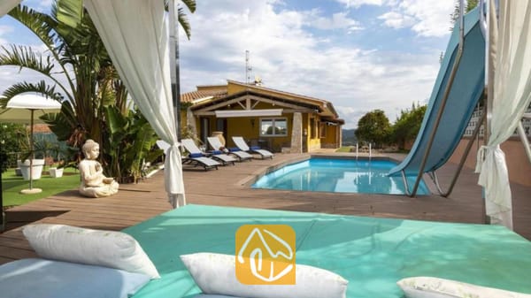 Vakantievilla's Costa Brava Spanje - Villa Soleia - Buitenzwembad en houten terras bij Villa Soleia, voorzien van ligstoelen, een blauwe waterglijbaan en een geel huis op de achtergrond.
