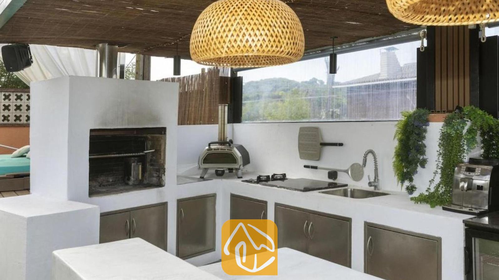 Vakantievilla's Costa Brava Spanje - Villa Soleia - Buitenkeuken bij Villa Soleia, voorzien van een ingebouwde barbecue, een pizzaoven, witte aanrechtbladen en geweven hanglampen.
