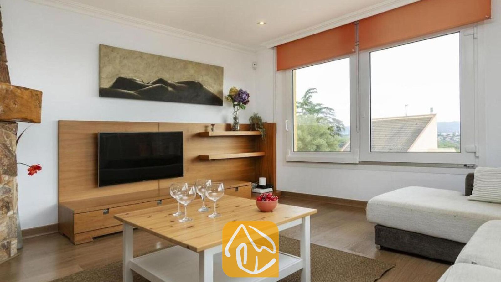 Vakantievilla's Costa Brava Spanje - Villa Soleia - Interieur van de woonkamer in Villa Soleia, met een houten salontafel voorzien van wijnglazen, een aan de muur bevestigde tv tegen houten panelen en grote ramen met oranje jaloezieën.