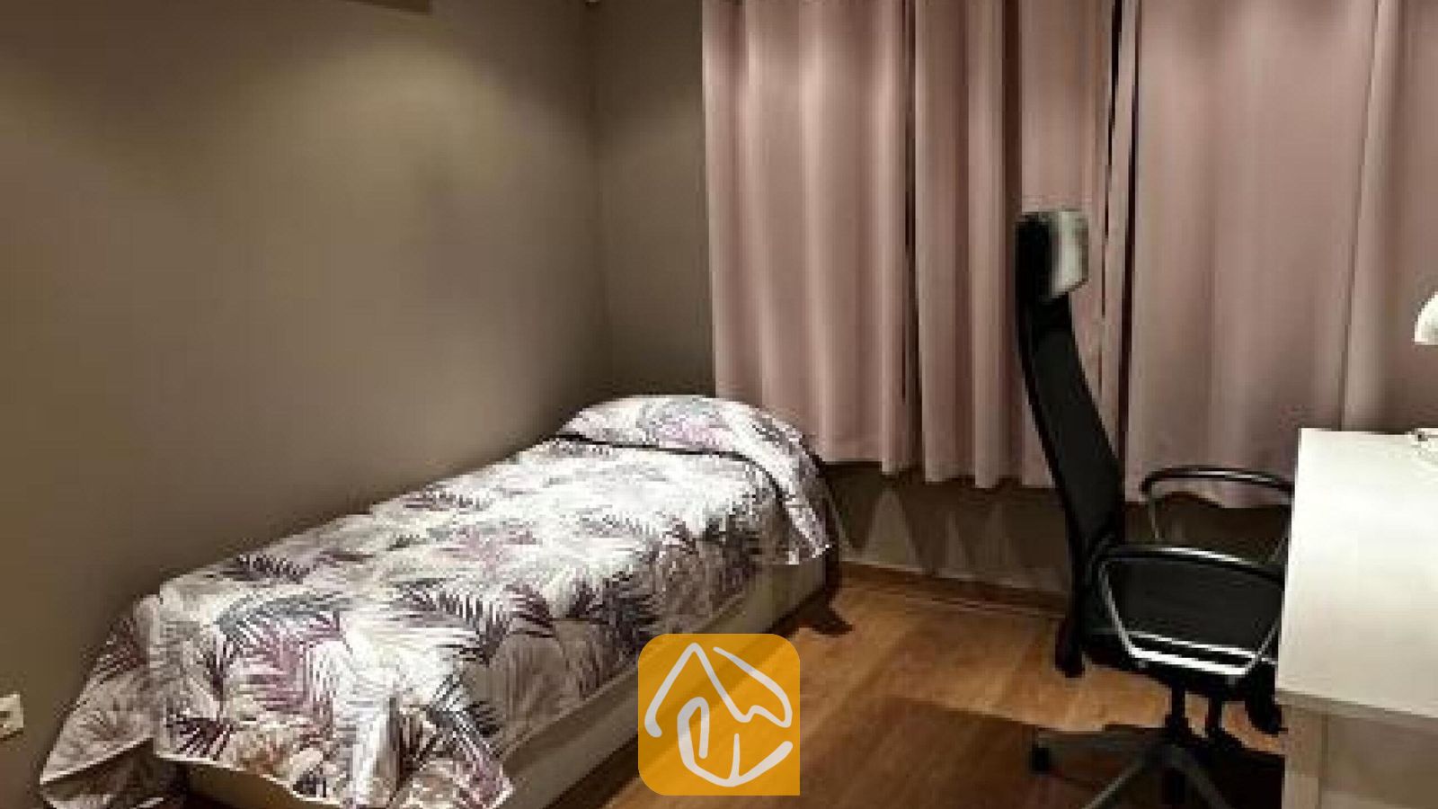 Vakantievilla's Costa Brava Spanje - Villa Soleia - Een éénpersoonsbed met een dekbed met botanisch motief naast een bureau en een zwarte bureaustoel in een slaapkamer met roze gordijnen.