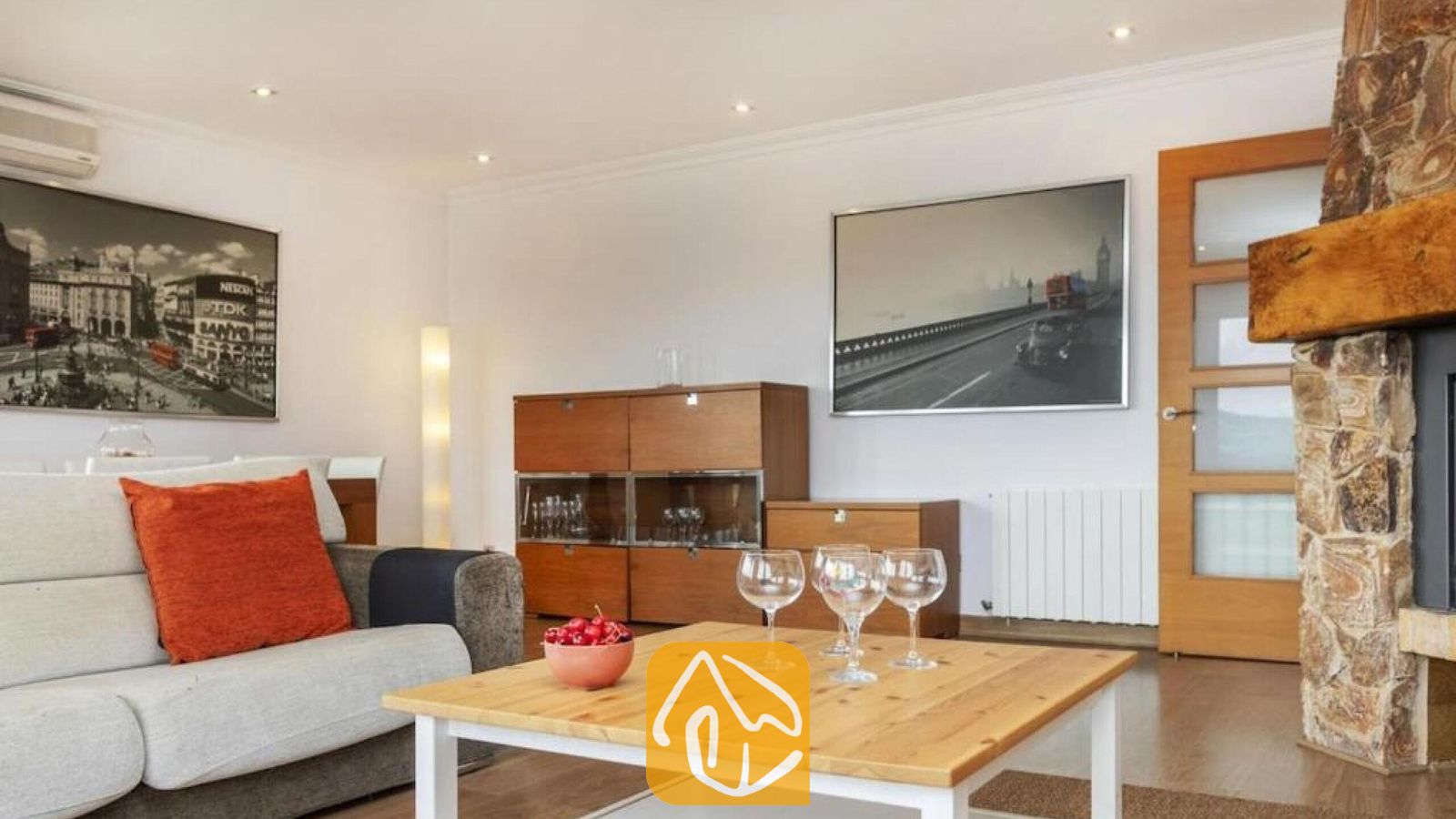 Vakantievilla's Costa Brava Spanje - Villa Soleia - Interieur van de woonkamer met een houten salontafel met wijnglazen, een lichtgrijze bank met een oranje kussen en een stenen open haard.