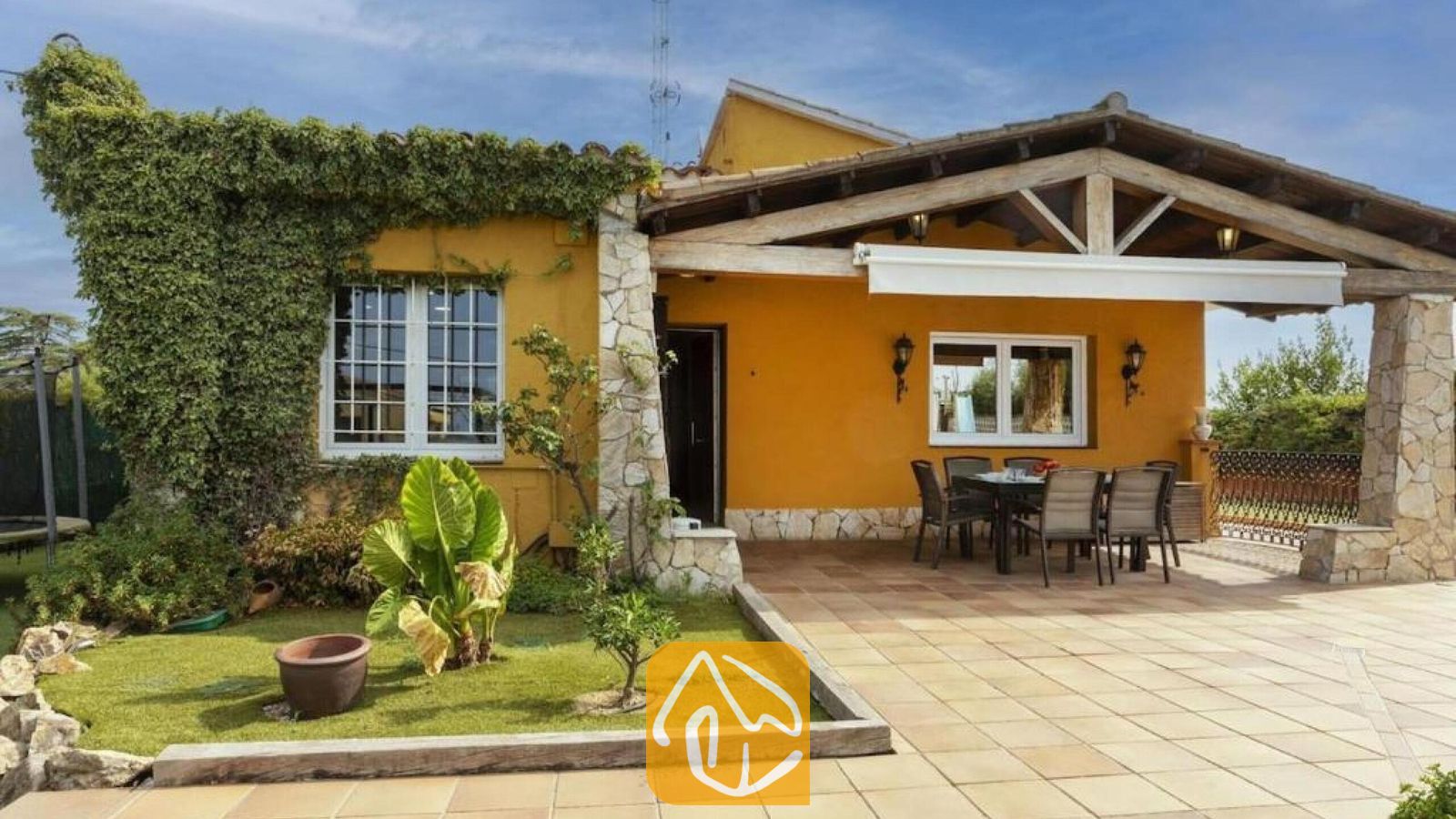Vakantievilla's Costa Brava Spanje - Villa Soleia - De gele buitengevel van Villa Soleia met een overdekt houten terras, een stenen terras en stenen pilaren naast een verzorgd gazon.