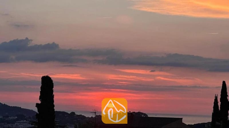 Villas de vacances Costa Brava Espagne - Villa Soleia - Un ciel de coucher de soleil aux nuances d'orange et de rose surplombant des collines lointaines, vu depuis la Villa Soleia avec des cyprès en ombres chinoises au premier plan.