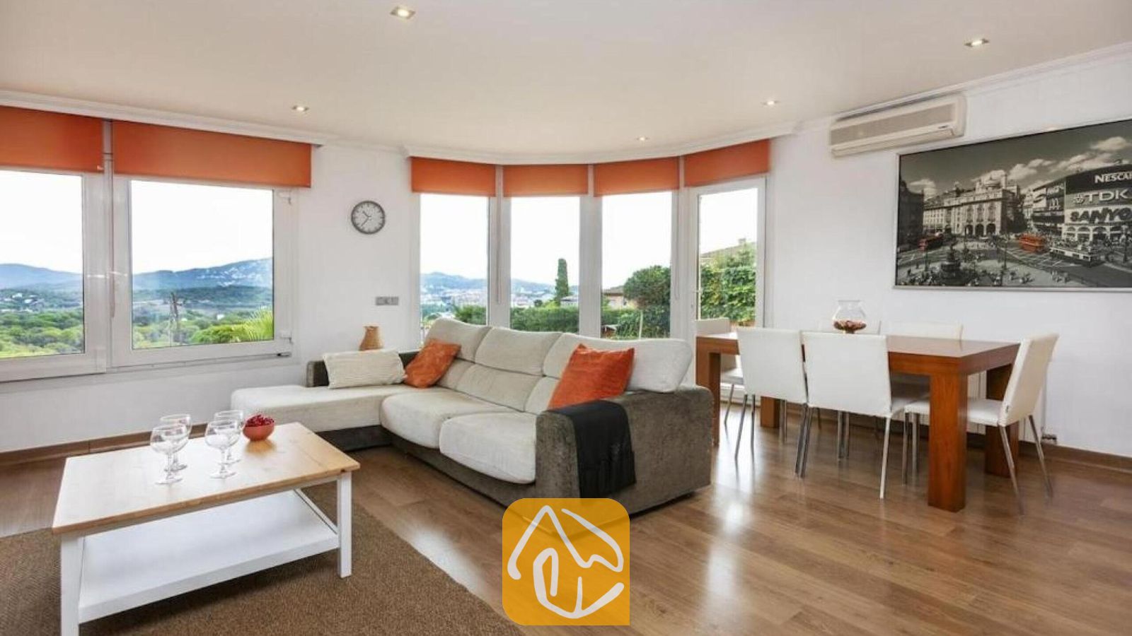 Vakantievilla's Costa Brava Spanje - Villa Soleia - Woon- en eetkamer van Villa Soleia met een lichtgrijs hoekbankstel, een houten salontafel en een eettafel met witte stoelen onder grote ramen met oranje jaloezieën.