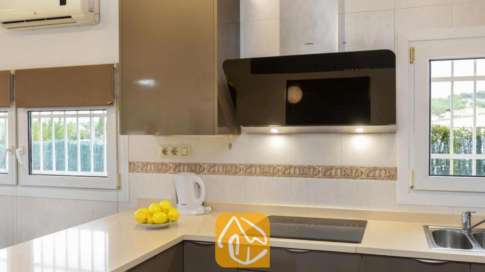 Vakantievilla's Costa Brava Spanje - Villa Soleia - Moderne keuken met beige aanrechtbladen, donkergrijze kasten, een zwarte glazen kookplaat en een schuine afzuigkap onder wandtegels met een decoratieve rand.