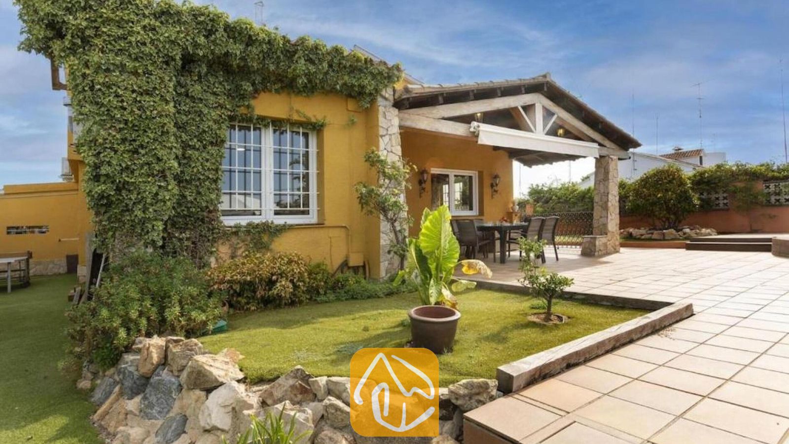 Vakantievilla's Costa Brava Spanje - Villa Soleia - Buitenaanzicht van Villa Soleia met gele stucwerkmuren begroeid met klimop, een overdekt stenen terras met een eethoek en een betegeld terras met een klein grasveld.