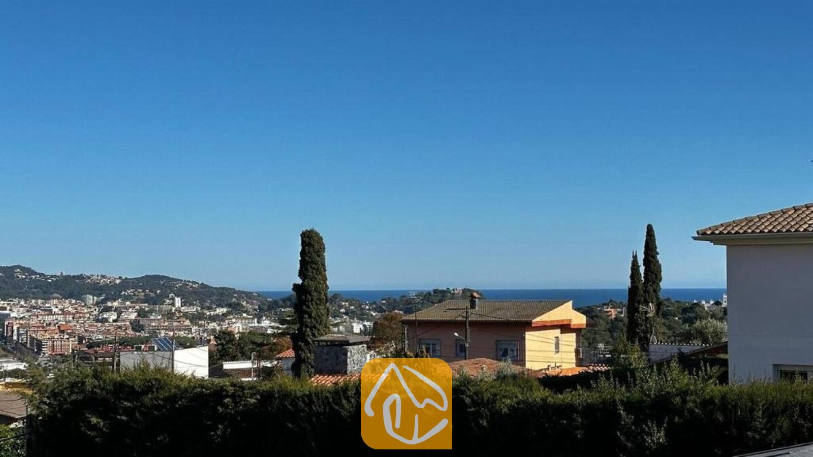 Vakantievilla's Costa Brava Spanje - Villa Soleia - Verheven uitzicht vanaf Villa Soleia met in de verte de gebouwen van het stadje, groene heuvels en een blauwe zeehorizon onder een heldere hemel.