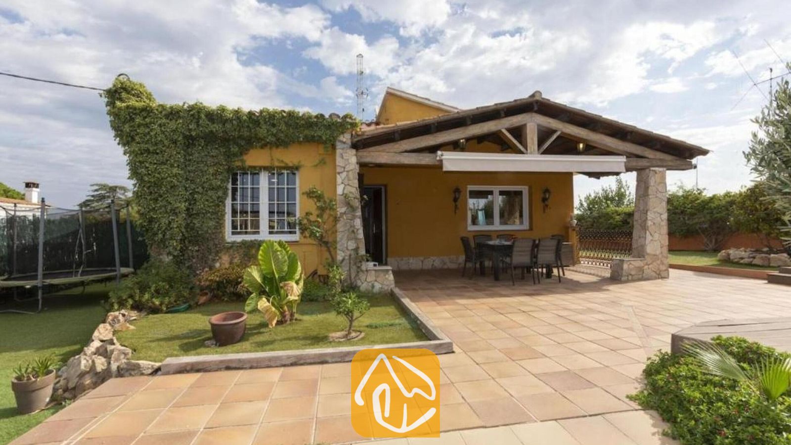 Vakantievilla's Costa Brava Spanje - Villa Soleia - Buitenaanzicht van Villa Soleia met een gele gevel begroeid met klimop, een overdekt terras met rode dakpannen en een eettafel, en stenen pilaren onder een houten balkenplafond.