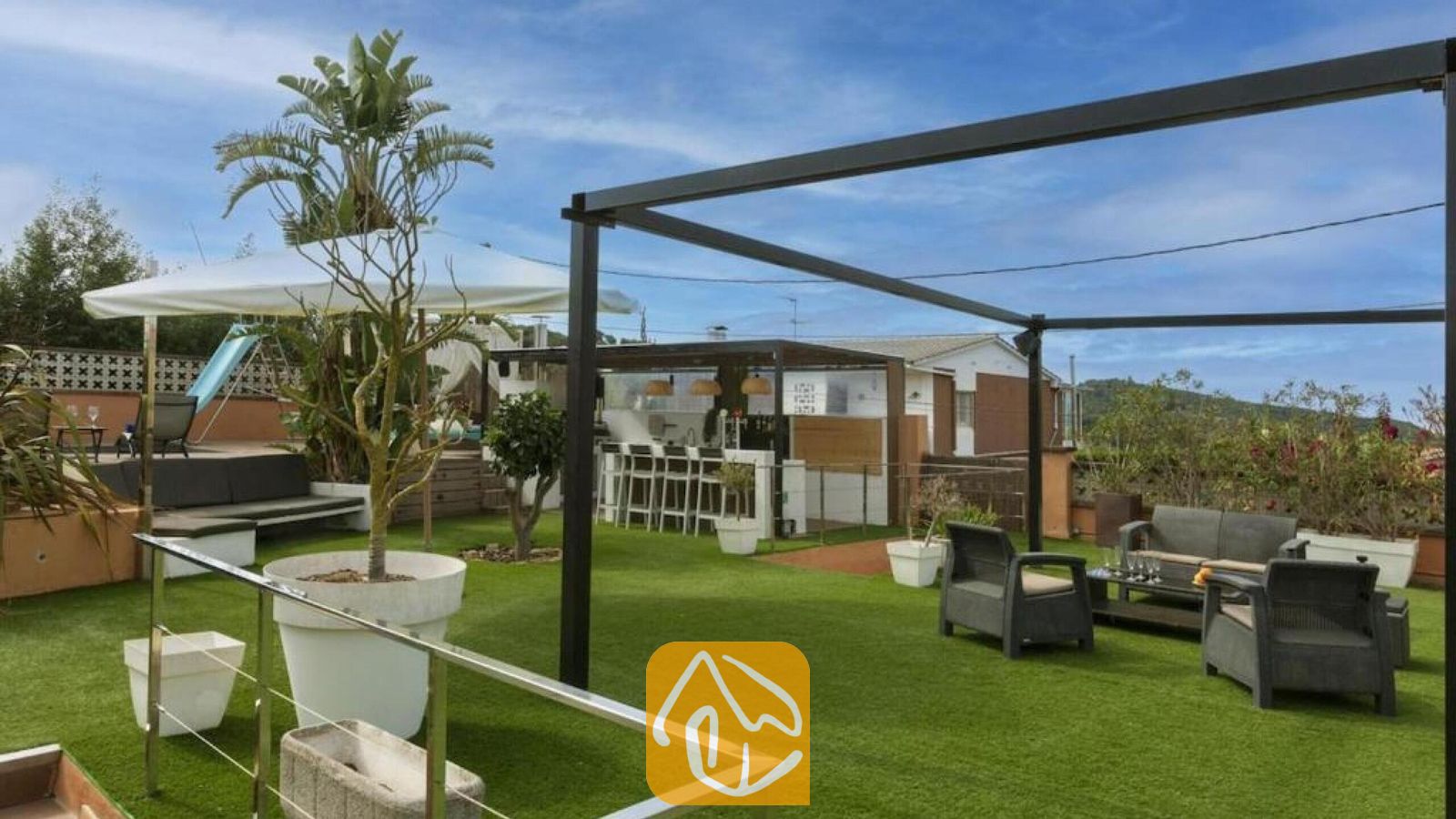 Vakantievilla's Costa Brava Spanje - Villa Soleia - Dakterras van Villa Soleia, voorzien van kunstgras, grijze fauteuils, een zwart metalen pergolaframes en een buitenbar.