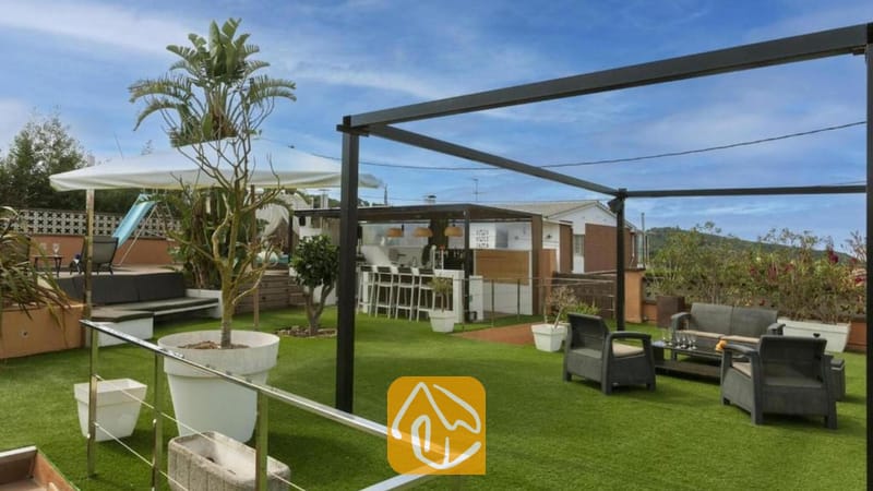 Vakantievilla's Costa Brava Spanje - Villa Soleia - Dakterras van Villa Soleia, voorzien van kunstgras, grijze fauteuils, een zwart metalen pergolaframes en een buitenbar.