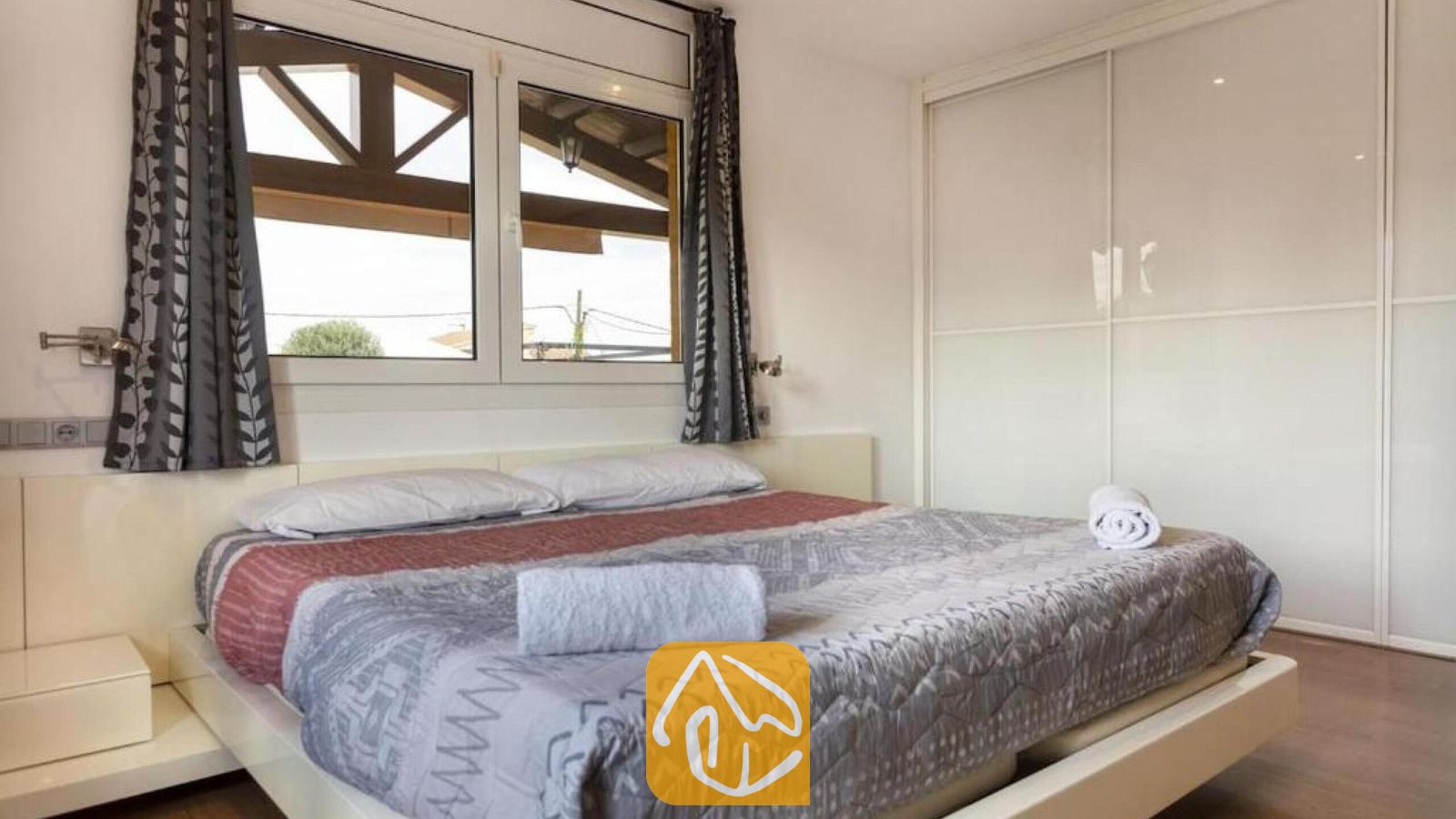 Vakantievilla's Costa Brava Spanje - Villa Soleia - Slaapkamer met een tweepersoonsbed opgemaakt met beddengoed in grijze en rode patronen, twee opgerolde witte handdoeken, witte kasten en ramen met grijze gordijnen met patroon.