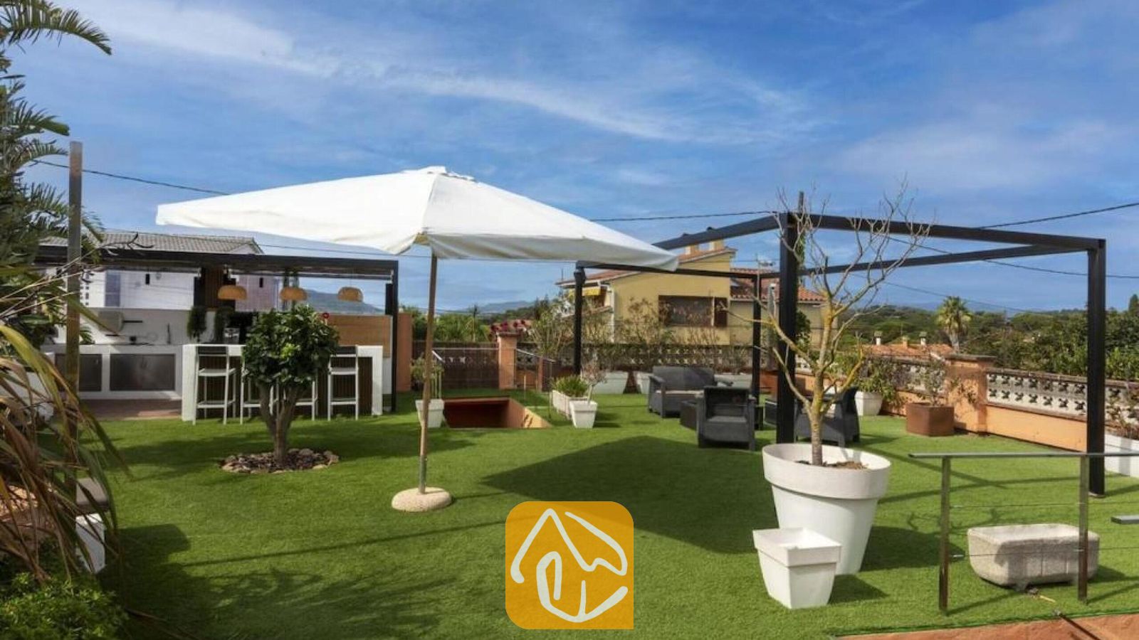 Vakantievilla's Costa Brava Spanje - Villa Soleia - Dakterras van Villa Soleia met groen kunstgras, een witte parasol, zwarte metalen pergola's, potplanten en zithoeken in de buitenlucht tegen een blauwe lucht.