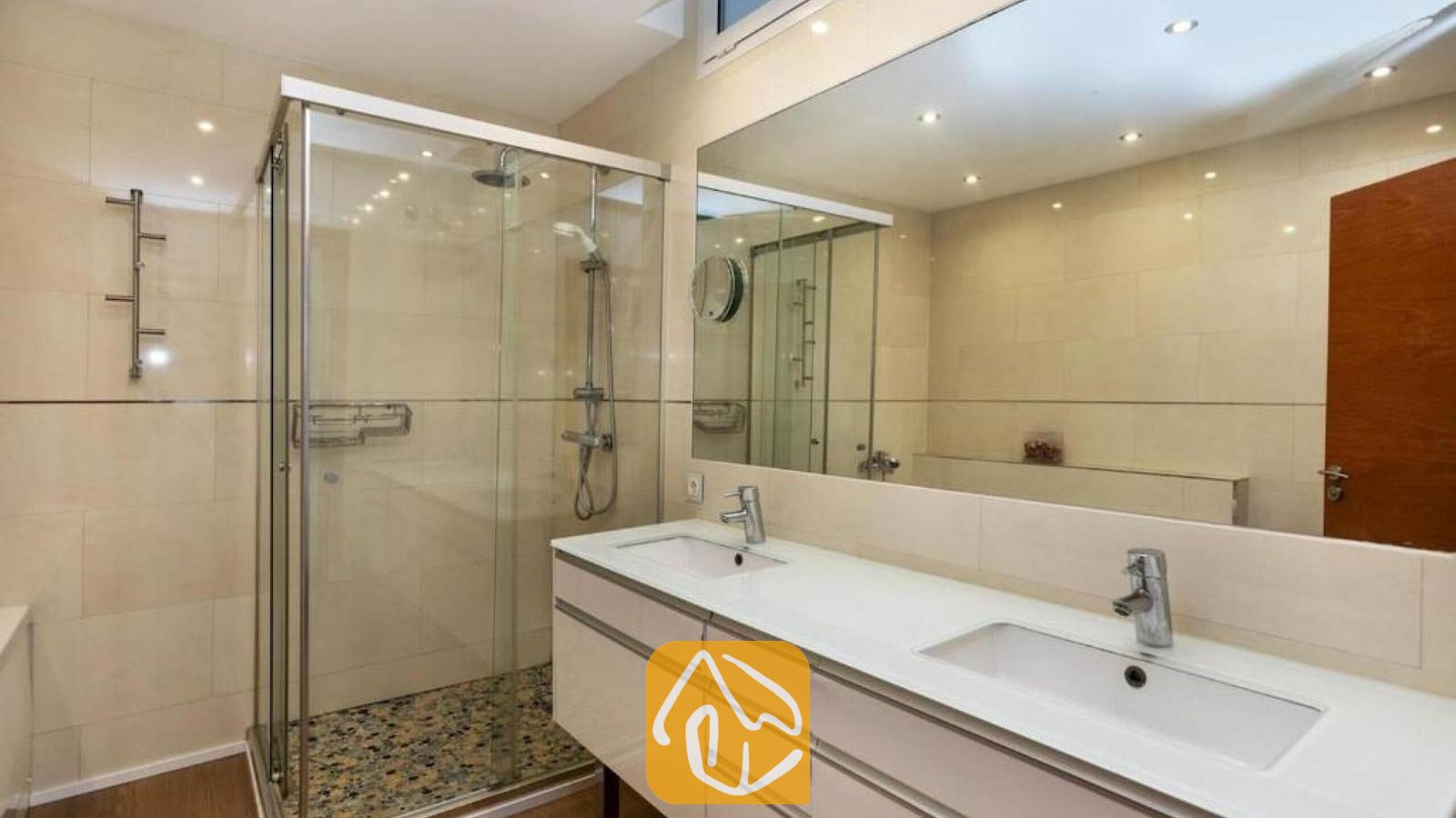 Vakantievilla's Costa Brava Spanje - Villa Soleia - Moderne badkamer voorzien van een dubbele wastafel met witte wasbakken, een grote wandspiegel en een glazen inloopdouche met een kiezeltegelvloer.