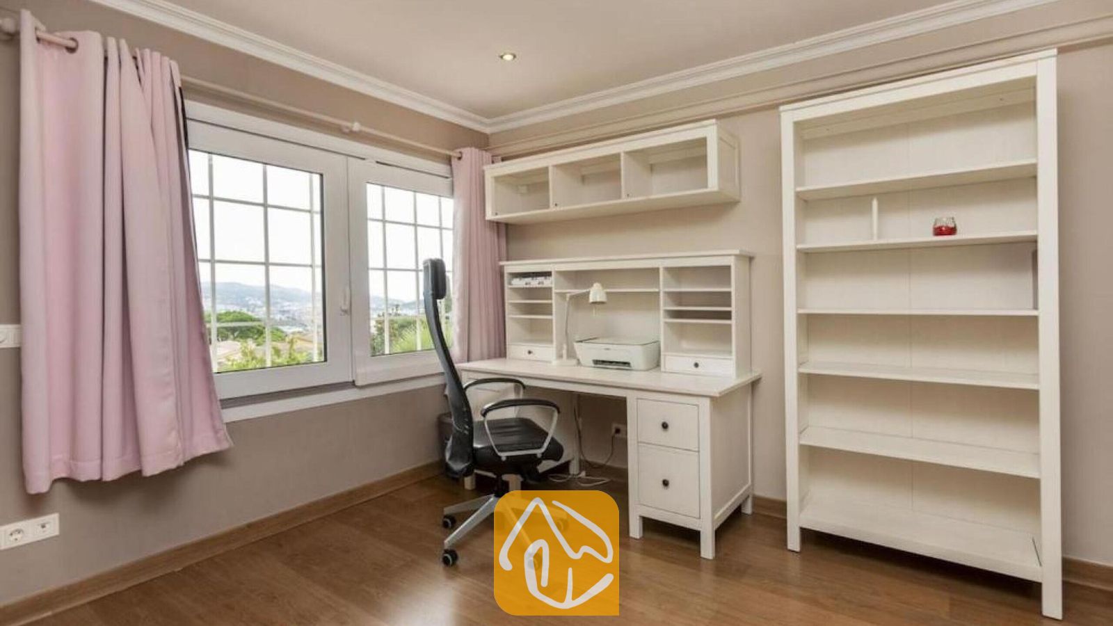 Vakantievilla's Costa Brava Spanje - Villa Soleia - Thuiskantoor met een wit bureau, een hoge boekenkast, een zwarte bureaustoel op wieltjes en een raam met roze gordijnen bij Villa Soleia.