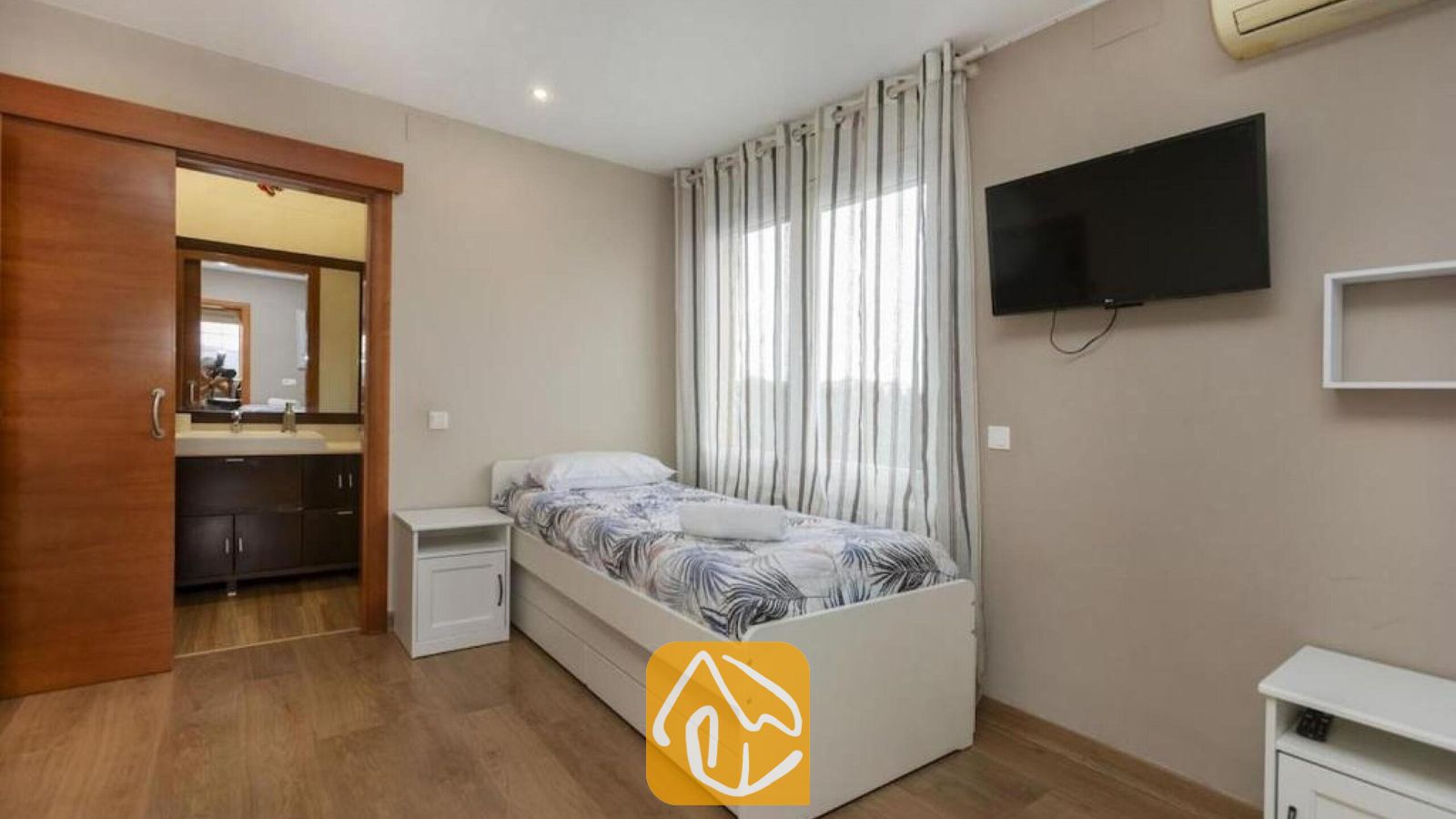 Vakantievilla's Costa Brava Spanje - Villa Soleia - Slaapkamer met een wit bed en beddengoed met motief, een wandgemonteerde flatscreen-tv en een aangrenzende badkamer met een donker badmeubel.