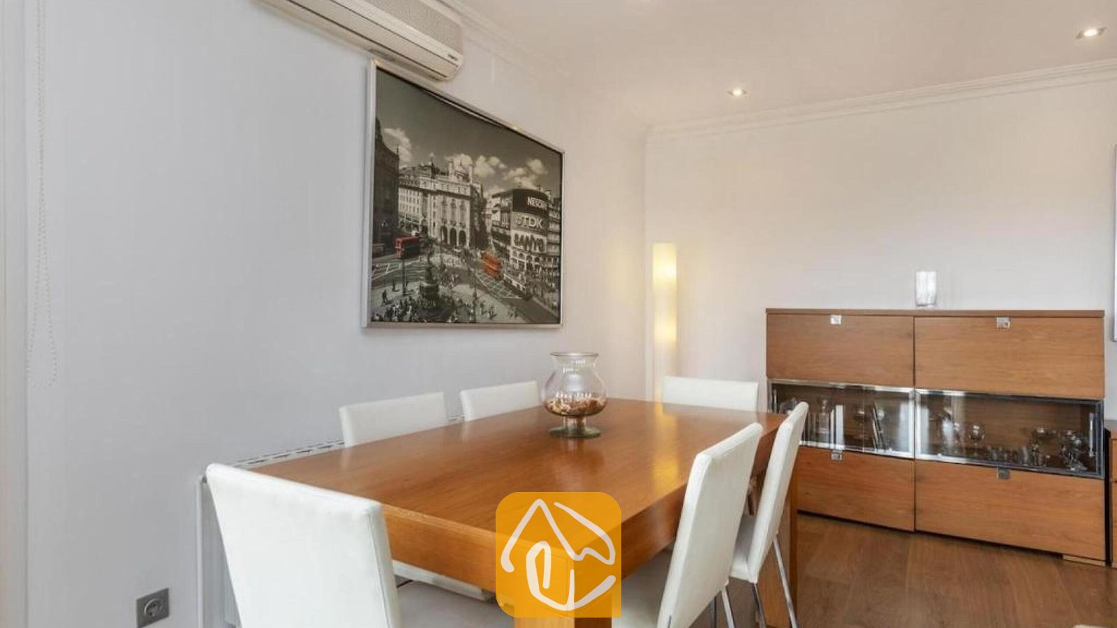 Vakantievilla's Costa Brava Spanje - Villa Soleia - Houten eettafel omringd door witte stoelen in Villa Soleia, voorzien van een groot ingelijst wandkunstwerk van een stadsgezicht en een houten opbergmeubel.