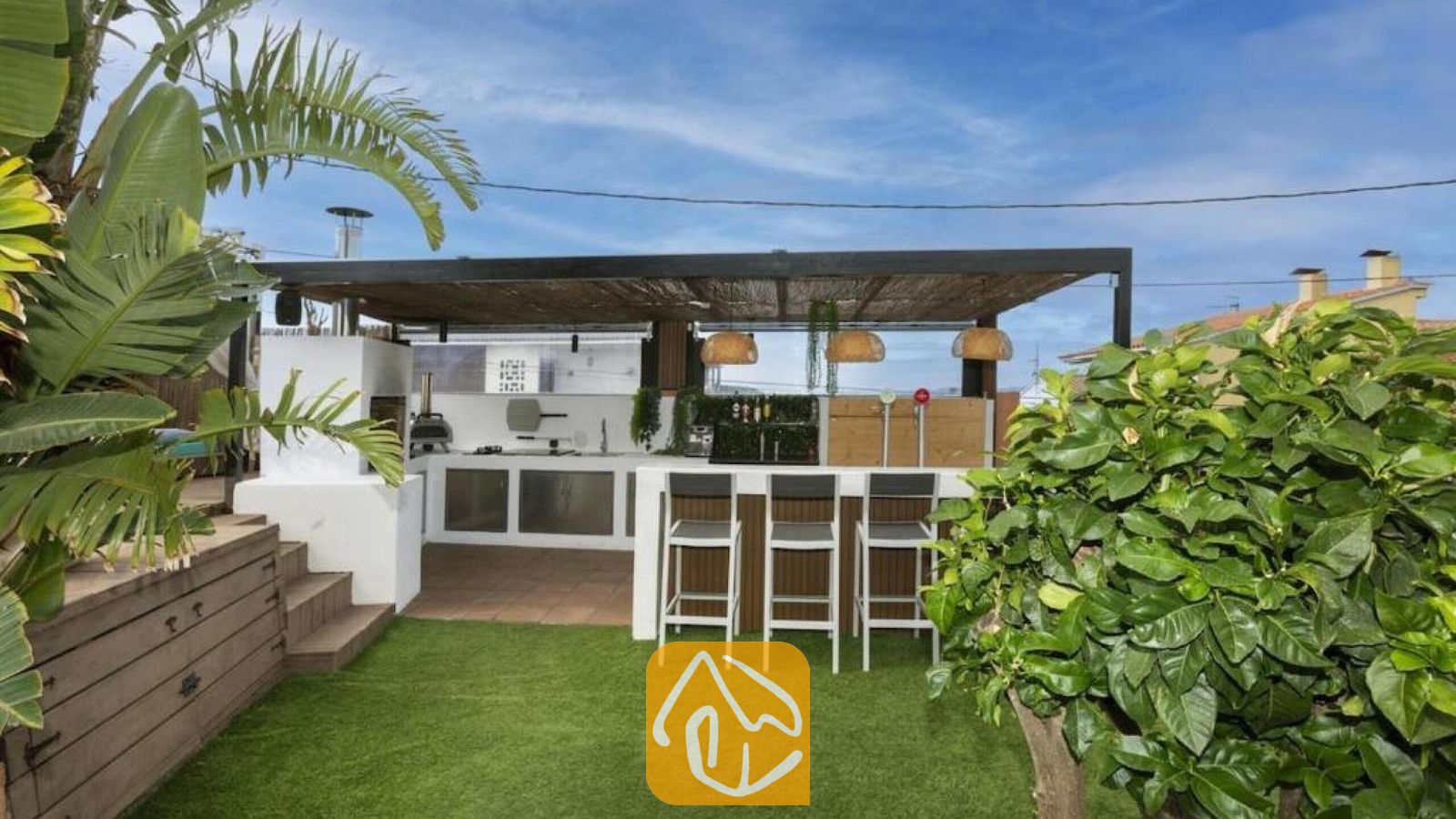 Vakantievilla's Costa Brava Spanje - Villa Soleia - Buitenkeuken en bar bij Villa Soleia, voorzien van een overdekte pergola, hoge barkrukken en een wit, ingebouwd kookeiland tegen een achtergrond van groen kunstgras.