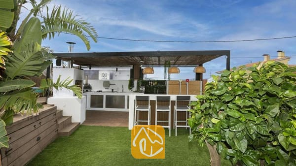 Vakantievilla's Costa Brava Spanje - Villa Soleia - Buitenkeuken en bar bij Villa Soleia, voorzien van een overdekte pergola, hoge barkrukken en een wit, ingebouwd kookeiland tegen een achtergrond van groen kunstgras.