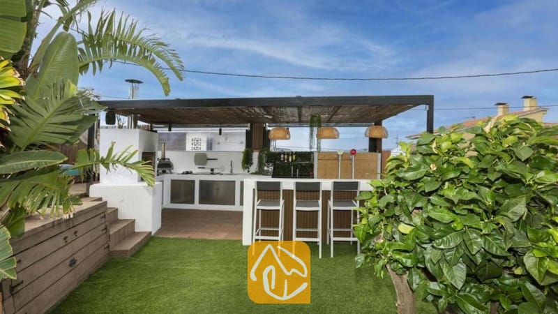 Vakantievilla's Costa Brava Spanje - Villa Soleia - Buitenkeuken en bar bij Villa Soleia, voorzien van een overdekte pergola, hoge barkrukken en een wit, ingebouwd kookeiland tegen een achtergrond van groen kunstgras.