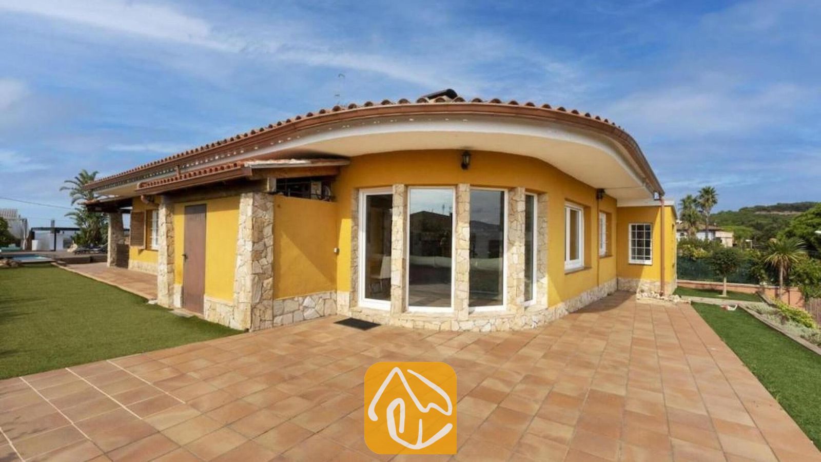 Vakantievilla's Costa Brava Spanje - Villa Soleia - Gele buitengevel van Villa Soleia met gebogen terracotta dakpannen, stenen zuilaccenten en een groot betegeld terras onder een blauwe lucht.