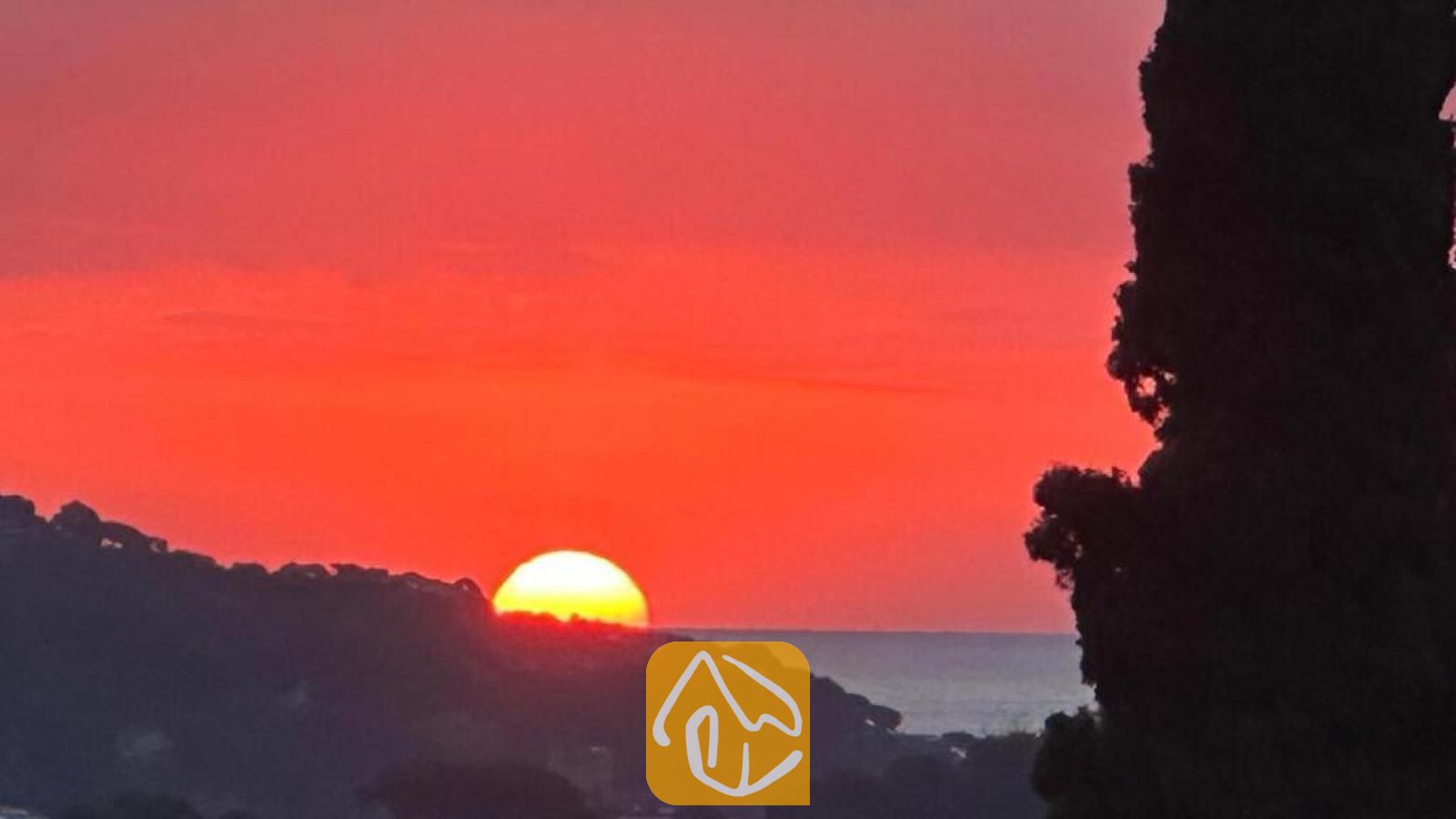 Vakantievilla's Costa Brava Spanje - Villa Soleia - Een levendige rood- en oranjekleurige zonsondergang boven verre heuvels en de zee, gezien vanuit Villa Soleia met het silhouet van een donkere boom op de voorgrond.