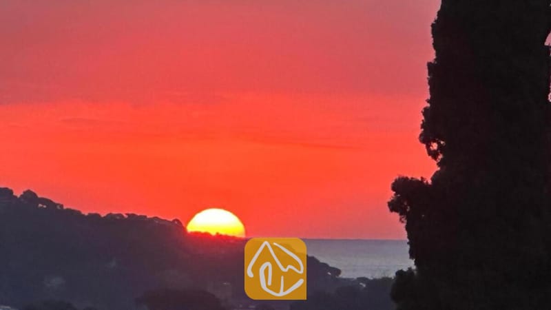 Villas de vacances Costa Brava Espagne - Villa Soleia - Coucher de soleil aux teintes vibrantes de rouge et d'orange sur les collines lointaines et la mer, admiré depuis la Villa Soleia, avec la silhouette sombre d'un arbre au premier plan.