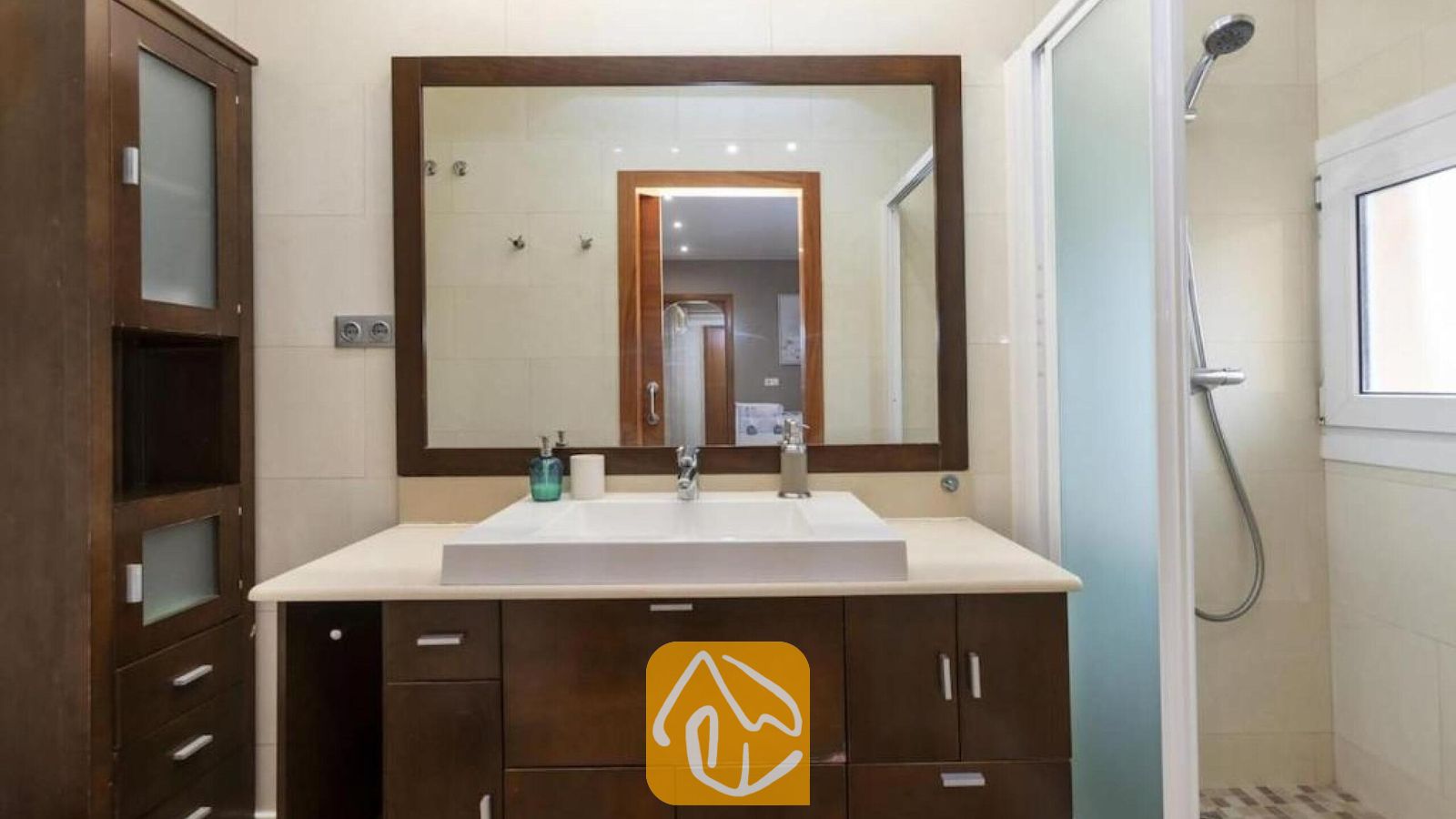 Vakantievilla's Costa Brava Spanje - Villa Soleia - Badkamermeubel met een donker houten kastje, een grote rechthoekige spiegel en een witte opzetwastafel naast een glazen douchecabine