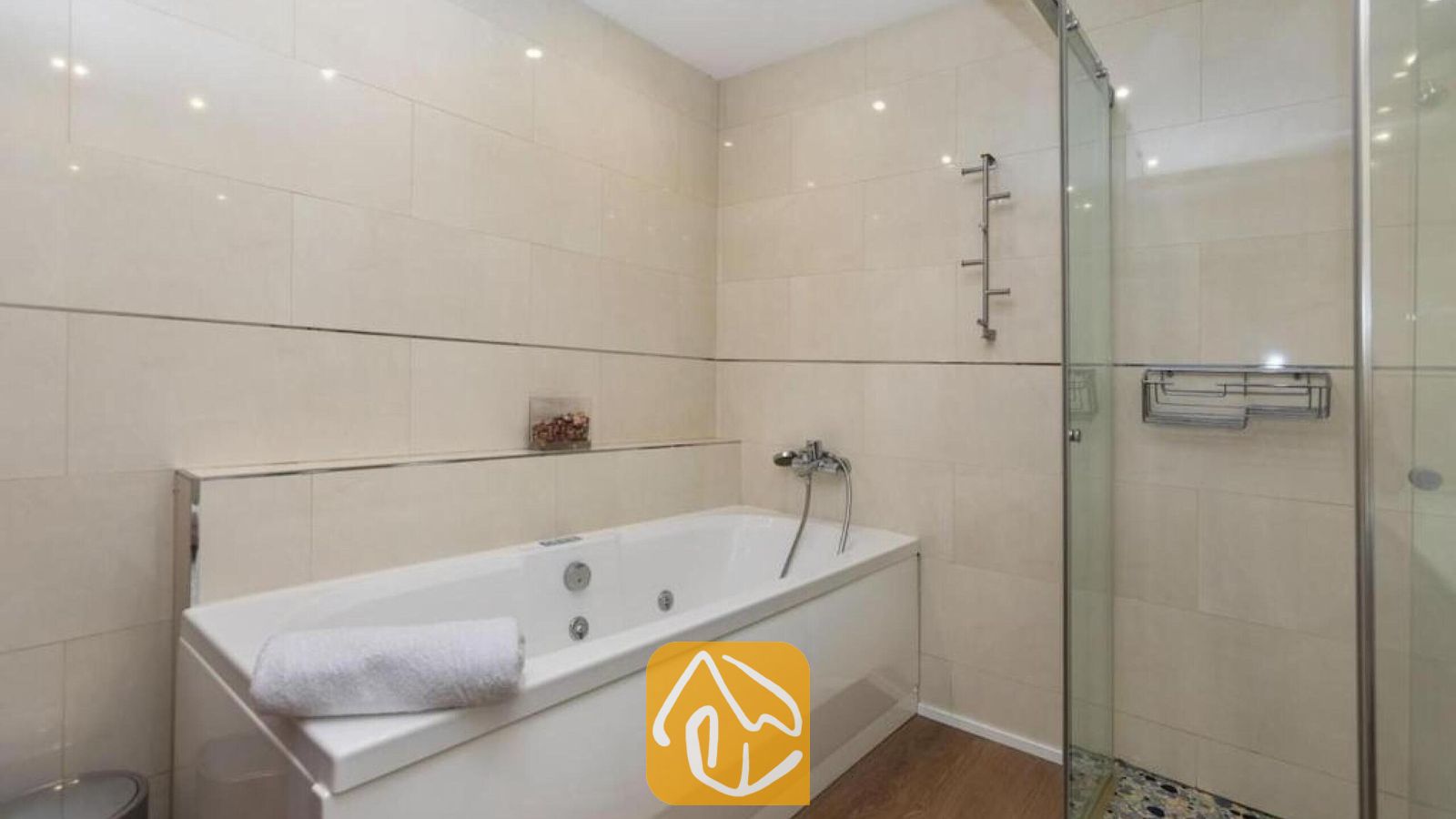 Vakantievilla's Costa Brava Spanje - Villa Soleia - Ligbad met een opgerolde witte handdoek, chroomkleurige armaturen en beige tegelwanden naast een glazen douchecabine