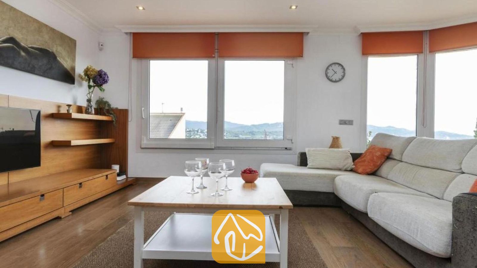 Vakantievilla's Costa Brava Spanje - Villa Soleia - Woonkamer van Villa Soleia, voorzien van een lichtgrijze hoekbank, een houten salontafel met wijnglazen en grote ramen met oranje rollo's die uitkijken op de verre heuvels.