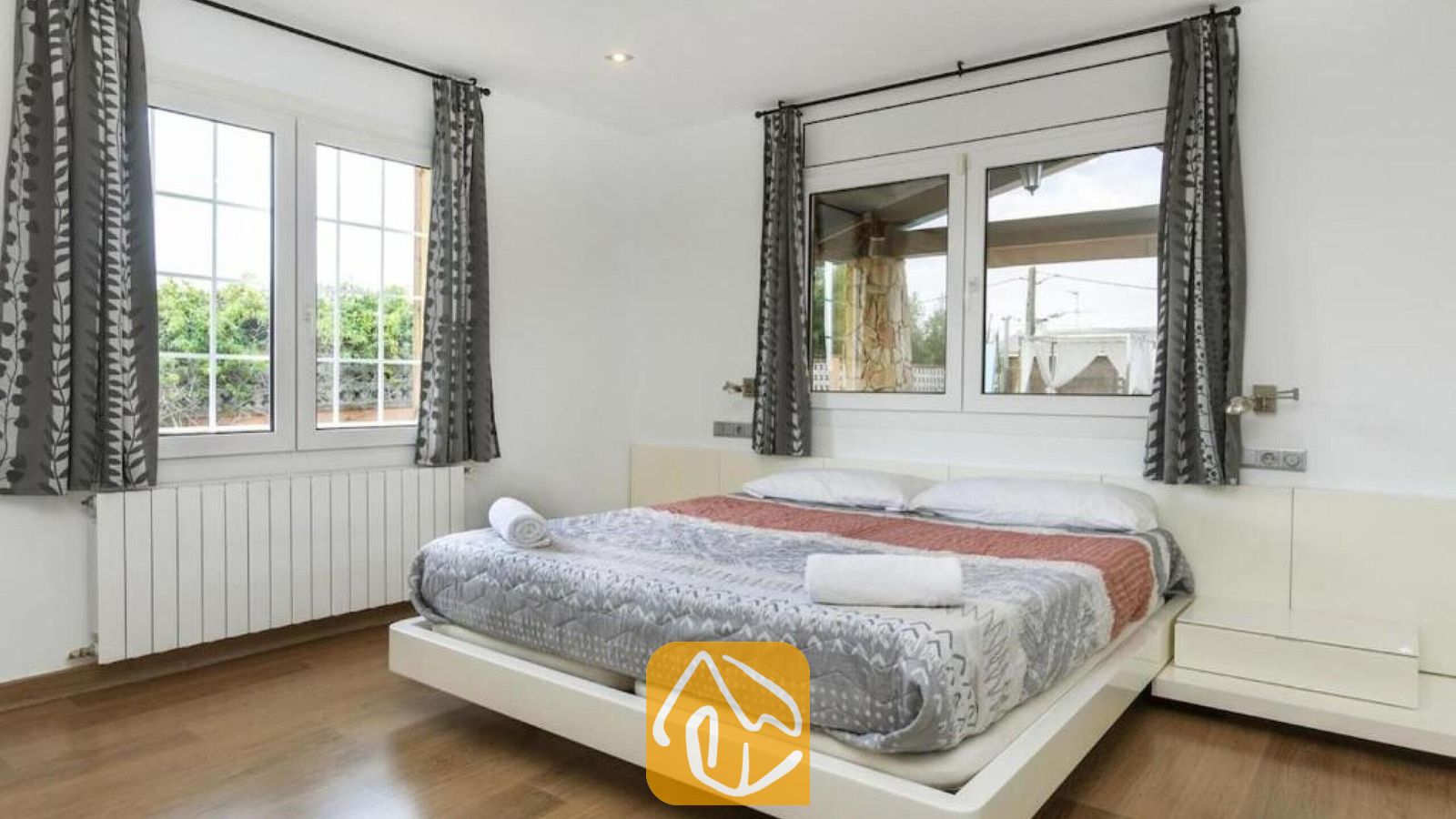 Vakantievilla's Costa Brava Spanje - Villa Soleia - Slaapkamer met een tweepersoonsbed, gedessineerd grijs en rood beddengoed, houten vloeren, witte muren en grote ramen met grijs gedessineerde gordijnen.