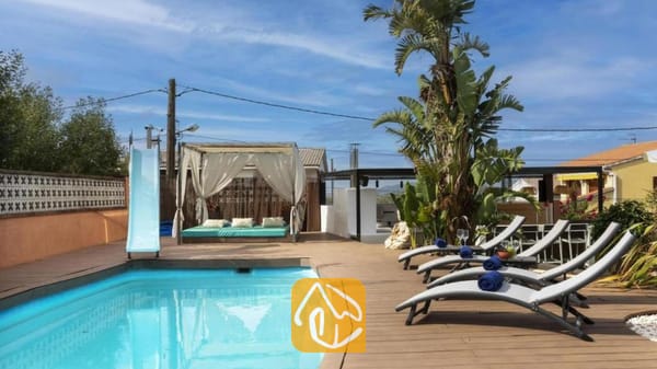 Vakantievilla's Costa Brava Spanje - Villa Soleia - Privézwembad met een blauwe glijbaan, houten terras, grijze ligstoelen met blauwe handdoeken en een gedrapeerde cabana bij Villa Soleia.