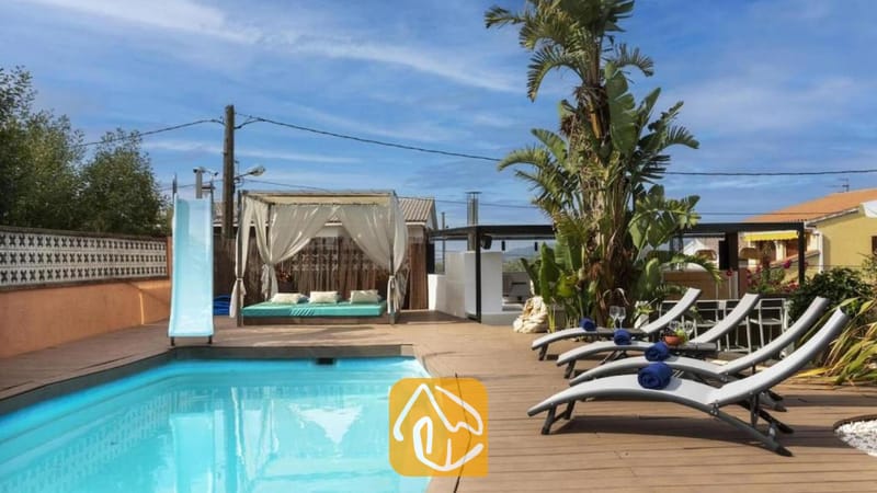 Vakantievilla's Costa Brava Spanje - Villa Soleia - Privézwembad met een blauwe glijbaan, houten terras, grijze ligstoelen met blauwe handdoeken en een gedrapeerde cabana bij Villa Soleia.