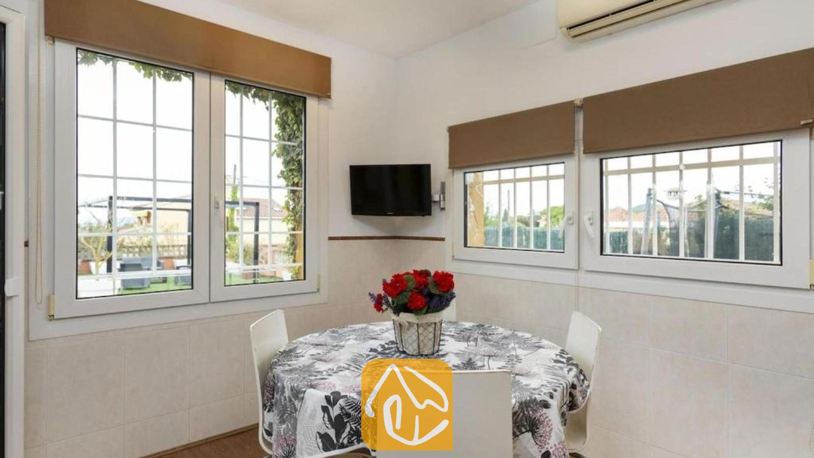 Vakantievilla's Costa Brava Spanje - Villa Soleia - Eethoek met een ronde tafel, een bloemendafelkleed, witte stoelen en een kleine televisie bevestigd aan de witgetegelde muur onder ramen met bruine rolgordijnen.
