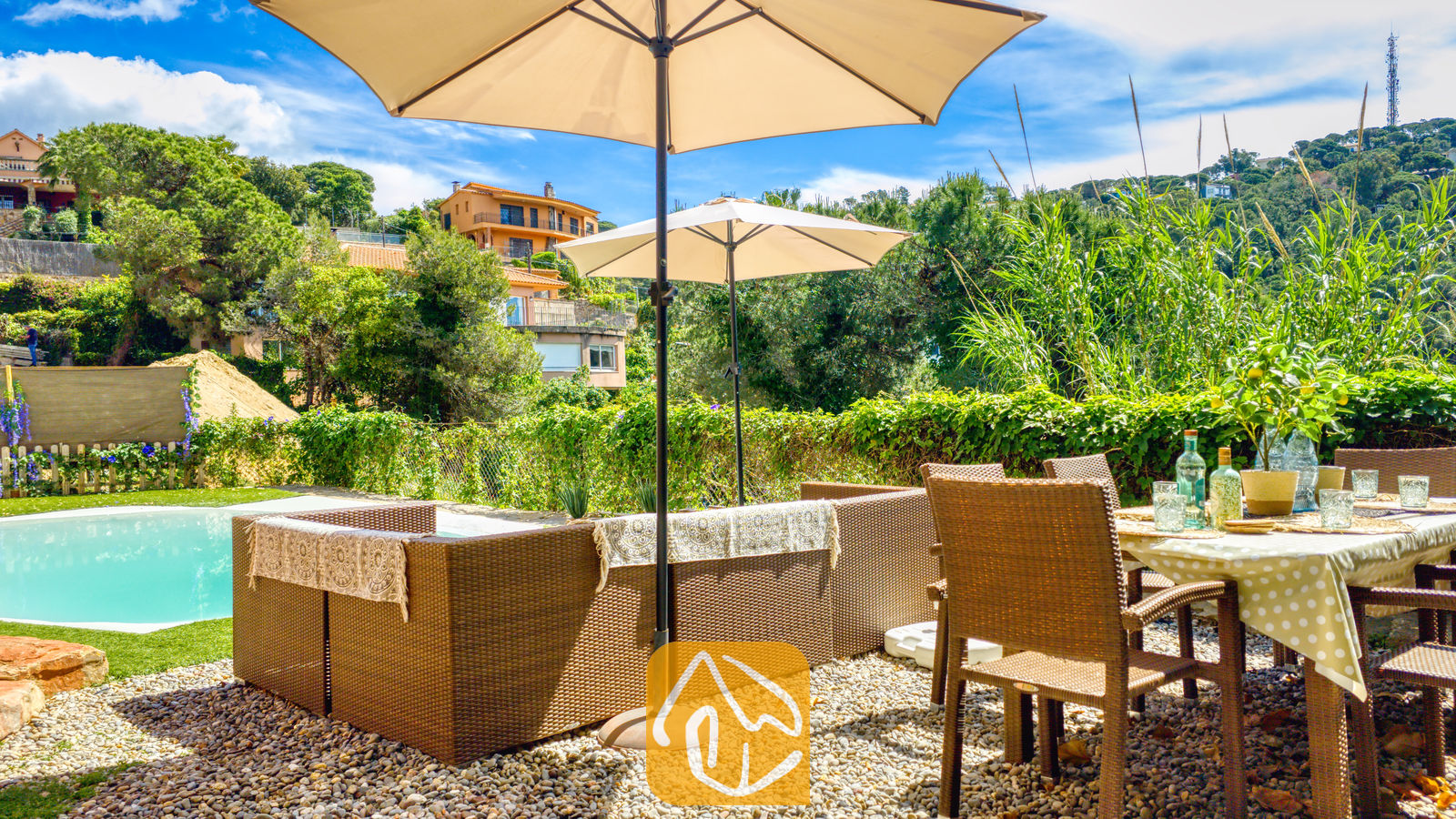 Vakantievilla's Costa Brava Spanje - Villa Sunny Beach - Buitenwinkeldinergedeelte en wicker loungewear-set onder beige parasols bij Villa Sunny Beach, voorzien van een grindterras, zwembad en een groene heuvelachtergrond.