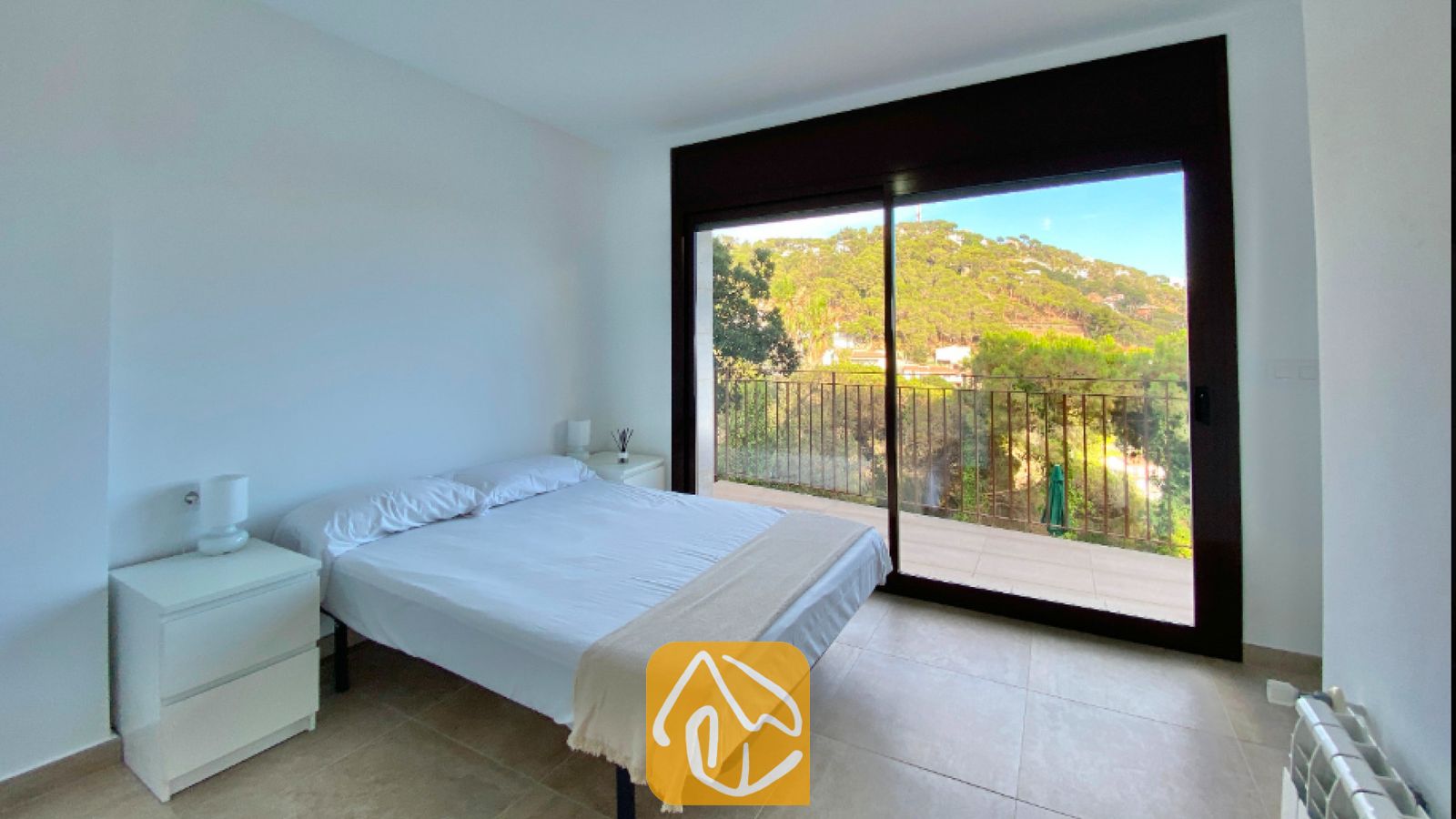 Vakantievilla's Costa Brava Spanje - Villa Sunny Beach - Slaapkamer met een wit tweepersoonsbed, witte nachtkastjes en een schuifpui die uitkomt op een balkon met uitzicht op groene heuvels.