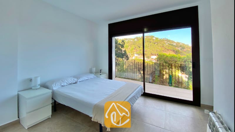 Villas de vacaciones Costa Brava España - Villa Sunny Beach - Dormitorio con una cama doble blanca, mesitas de noche blancas y puertas corredizas de cristal que se abren a un balcón con vistas a verdes colinas.