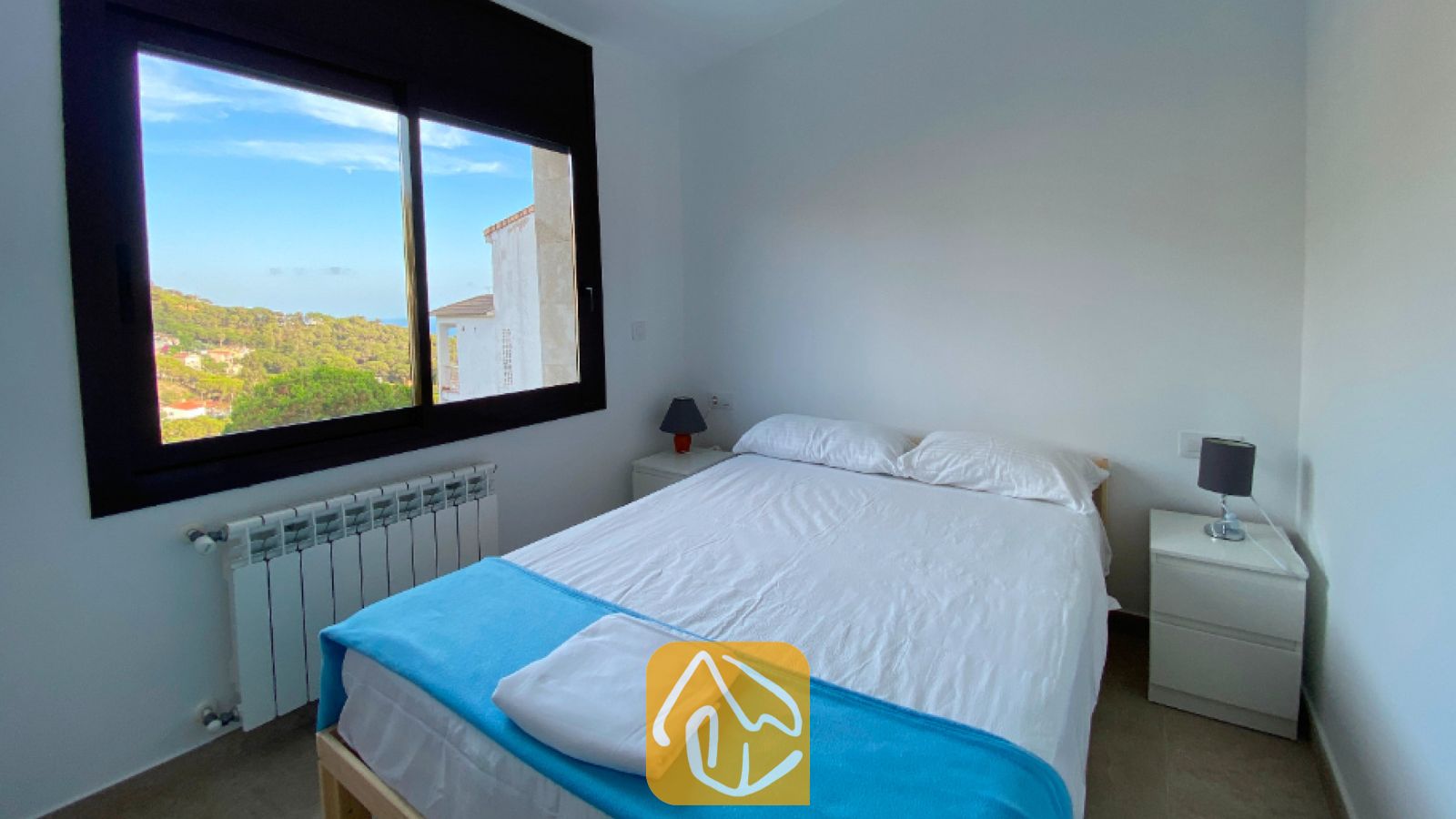 Vakantievilla's Costa Brava Spanje - Villa Sunny Beach - Slaapkamer met een tweepersoonsbed opgemaakt in wit linnen en voorzien van een blauwe sprei, geflankeerd door witte nachtkastjes met donkere lampen, en een raam dat uitzicht biedt op een heuvellandschap.