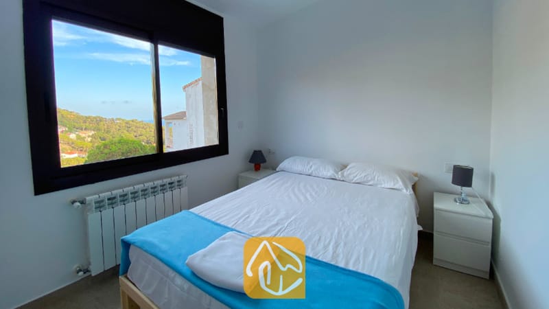 Villas de vacaciones Costa Brava España - Villa Sunny Beach - Dormitorio con cama doble vestida con ropa de cama blanca y una manta decorativa azul, flanqueada por mesitas de noche blancas con lámparas oscuras, y una ventana que enmarca una vista a la colina.