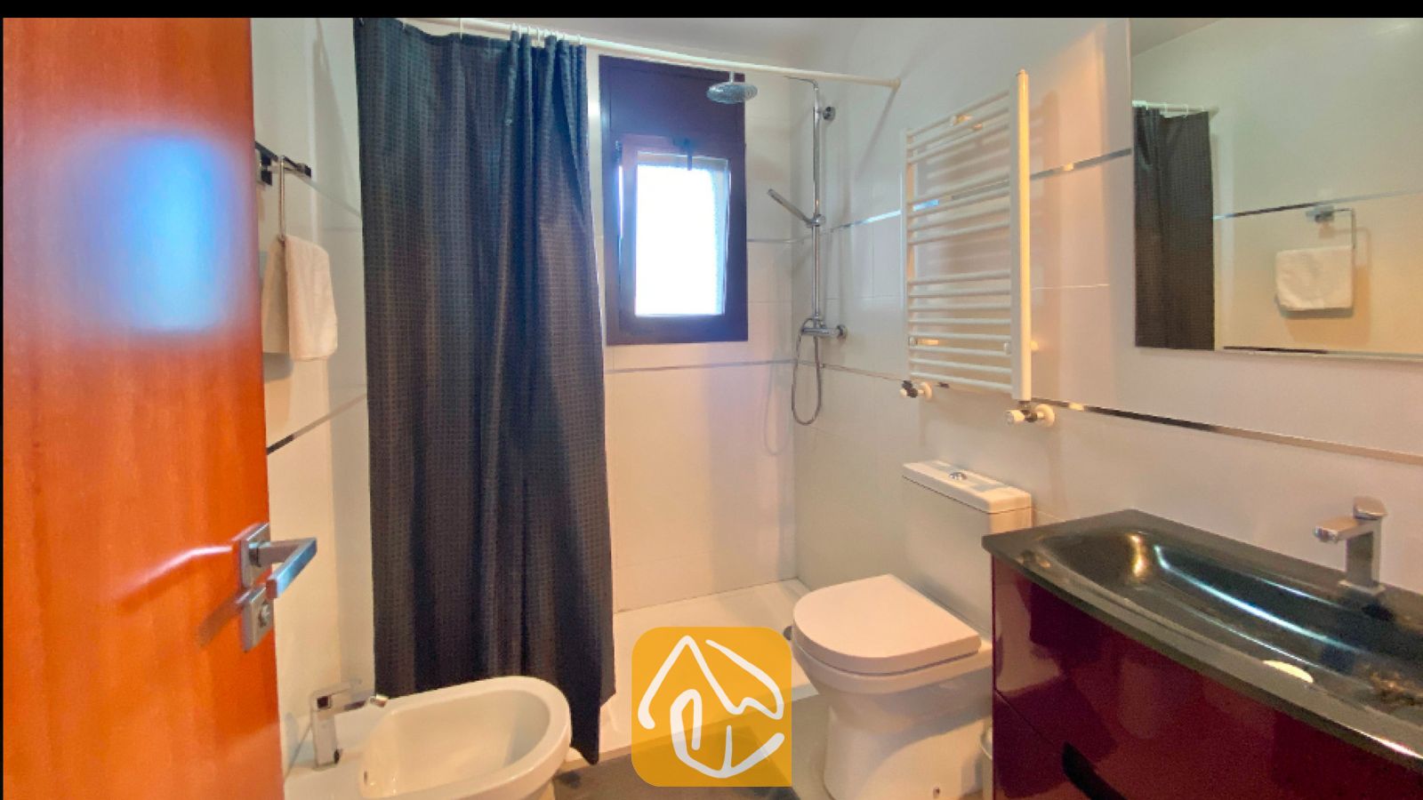 Vakantievilla's Costa Brava Spanje - Villa Sunny Beach - Badkamer voorzien van een donkergrijze douchegordijn, witte tegelmuren, een wit toilet en een roodbruin badkamermeubel met een zwarte wasbak.
