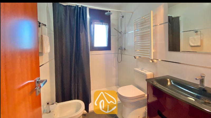 Villas de vacaciones Costa Brava España - Villa Sunny Beach - Baño equipado con una cortina de ducha en gris oscuro, paredes revestidas de azulejos blancos, inodoro blanco y un mueble de lavabo en tono marrón rojizo con lavabo negro.