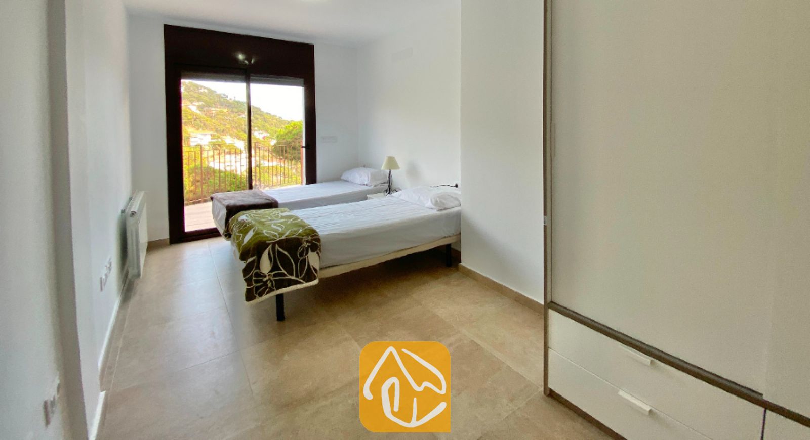 Vakantievilla's Costa Brava Spanje - Villa Sunny Beach - Slaapkamer met twee eenpersoonsbedden, wittewanden, tegelvloer, een grote kledingkast en glazen schuifdeuren die openen naar een uitzicht op de heuvels.