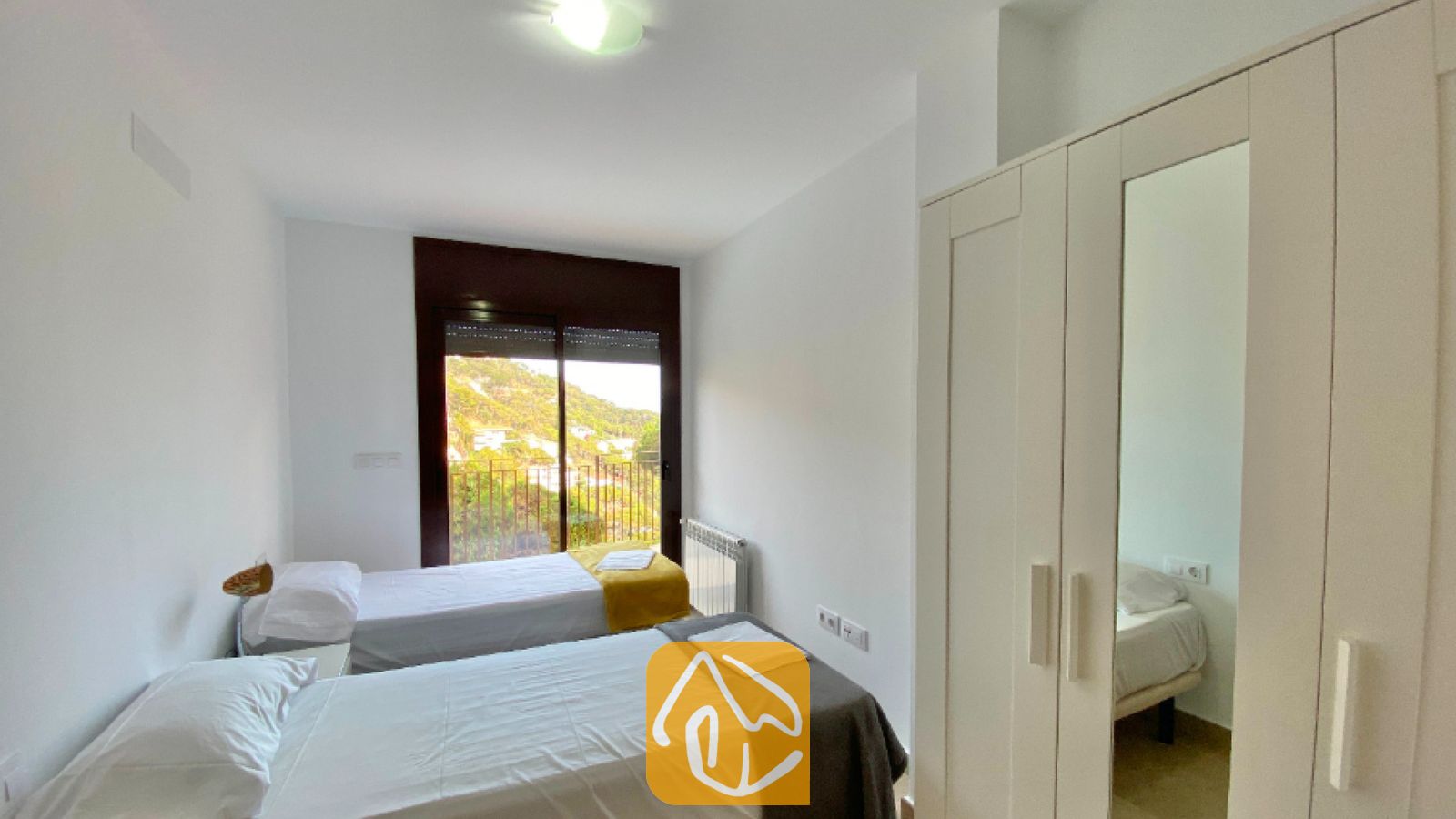 Vakantievilla's Costa Brava Spanje - Villa Sunny Beach - Slaapkamer met twee eenpersoonsbedden opgemaakt met wit beddengoed, een grote witte kledingkast met een gespiegelde deur en glazen schuifpui die toegang biedt tot een balkon met uitzicht op de heuvels.