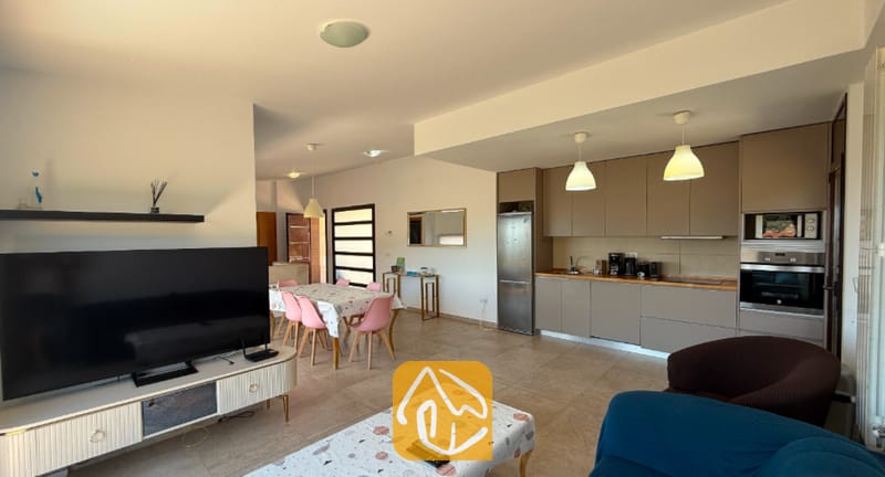 Vakantievilla's Costa Brava Spanje - Villa Sunny Beach - Open woonkamer met een grote flatscreen-tv, tegelvloeren, een eettafel met roze stoelen en een aangrenzende keuken met beige kasten.
