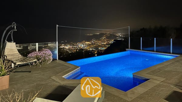 Vakantievilla's Costa Brava Spanje - Villa VistaMar - Verlicht zwembad bij Villa VistaMar in de nacht, met tegelvloeren, een hangstoel en een ver uitzicht op de lichtjes van de kuststad.