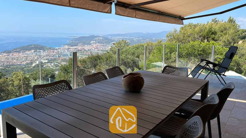 Vakantievilla's Costa Brava Spanje - Villa VistaMar - Overdekt buitenbuitenterras bij Villa VistaMar met een donkere houten tafel, gaasstoelen en een panoramisch uitzicht op zee en de kust.
